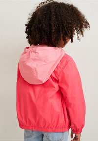 Bambino con i capelli ricci che indossa una giacca rosa brillante con cappuccio rosa chiaro, in piedi rivolto all'indietro su uno sfondo semplice.