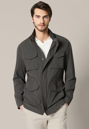 Młody mężczyzna w ciemnoszarej kurtce z czterema patkami kieszeniowymi na białej koszuli, stojący z rękami w kieszeniach jasnych spodni.