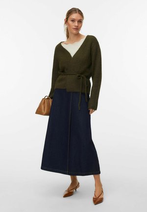 Femme portant un pull cache-cœur vert, un top blanc, une longue jupe sombre, des talons pointus marron, et tenant un petit sac à main marron.