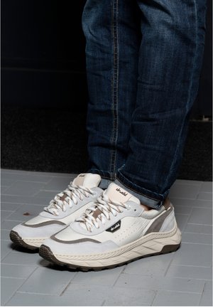 Person wearing white and gray casual sneakers with patterned laces and blue jeans, standing on gray tiled floor.