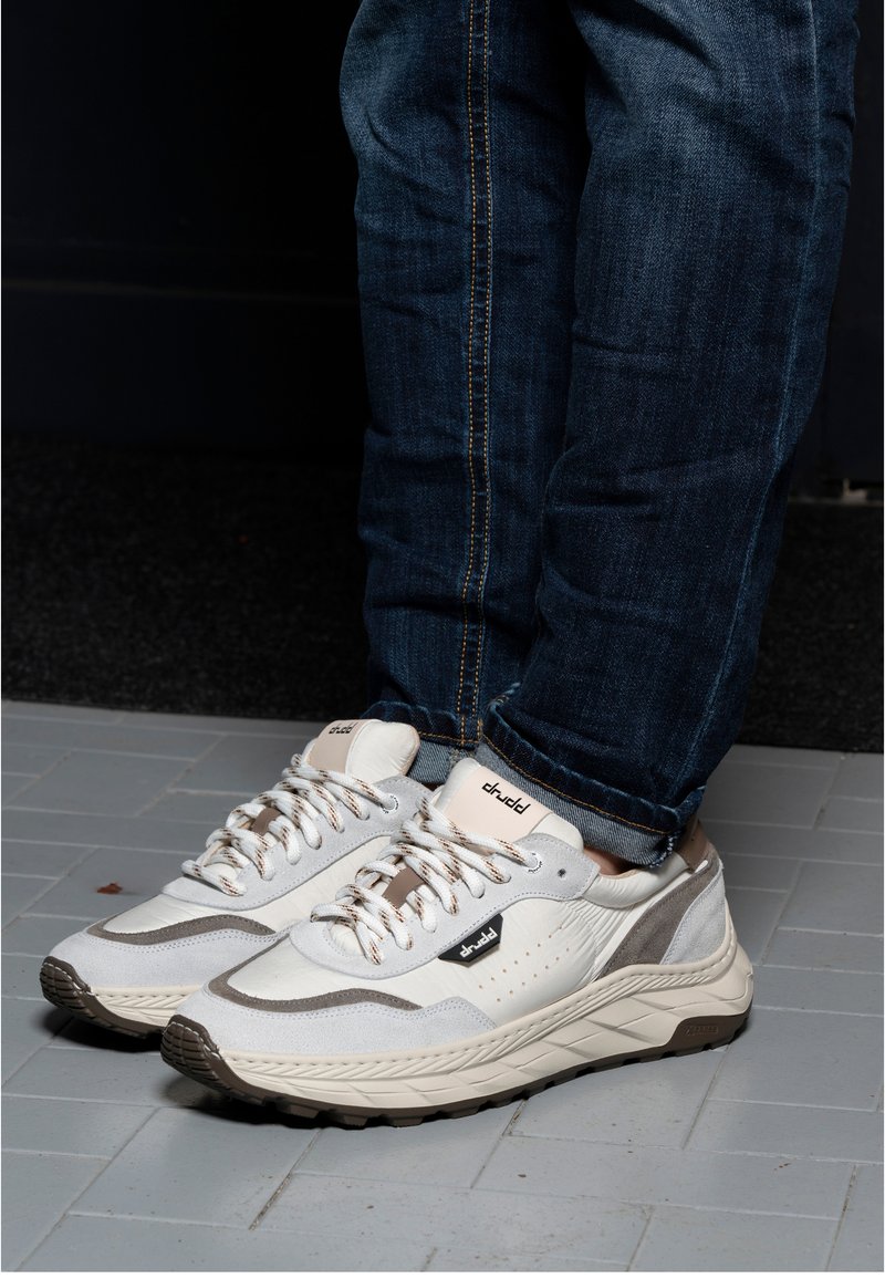 Persoon draagt witte en grijze casual sneakers met patroonveters en blauwe jeans, staand op een grijze tegelvloer.
