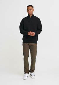 Svart genser med glidelås i mykt strikket materiale, kombinert med olivengrønne bukser og hvite sneakers med svarte striper. Enkelt design med ribbestrikkede detaljer.
