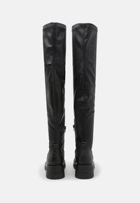 Mustat nahkaiset polven ylle ulottuvat saappaat, joissa on teksturoitu pinta. Niissä on pyöreä kärki, painava pohja, koko pituudelta avautuvat sivuvetoketjut ja ompeluyksityiskohtia.