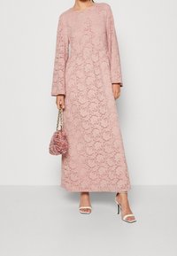 Różowa koronkowa sukienka maxi z długimi rękawami, kwiatowym wzorem i okrągłym dekoltem. Połączona z teksturowaną różową torebką i białymi szpilkami.