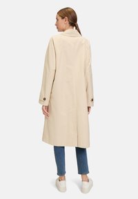 Cappotto trench beige con una vestibilità rilassata, caratterizzato da un colletto classico, maniche lunghe e polsini con bottone, abbinato a jeans blu e sneaker bianche.