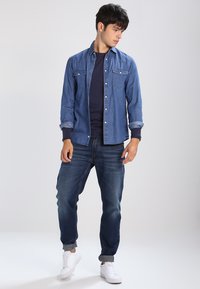 Denim shirt in light blue with two chest pockets, paired with a navy crew-neck top, dark blue jeans, and white sneakers. Casual fit.