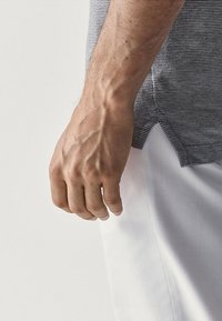 Main gauche veineuse détendue à côté du corps, portant une chemise grise texturée et un pantalon blanc, sur fond uni.