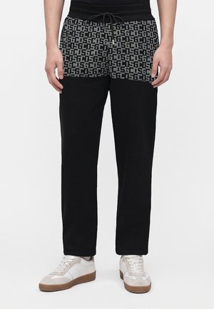 Personne portant un pantalon noir avec une ceinture grise et noire à motif et des baskets blanches avec des semelles marron clair, debout sur un fond blanc.