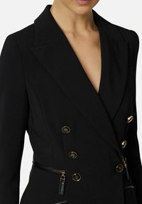 Blazer nero su misura con scollo a V profondo, design doppiopetto, bottoni dorati e dettaglio con cerniera in vita. Tessuto liscio e strutturato.