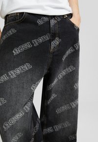 Svarta denimjeans med upprepat "NEW YORK"-grafik i ljusgrått. Har orange sömmar och en klassisk femfickadesign.