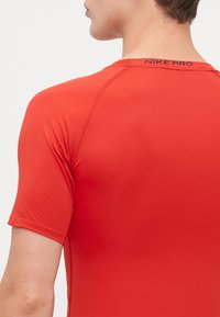Cămașă roșie Nike Pro cu maneci scurte, având o textură netedă și elastică, cu cusături de umăr întărite și un logo mic la guler.
