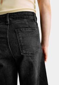 Zwart hoge taille denim broek met een achterzak, zichtbare stiknaad en een relaxte pasvorm, getoond vanuit de achterkant met een beige shirt in beeld.