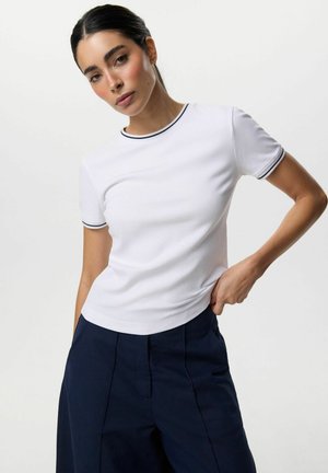 Kobieta o ciemnych włosach ubrana w białą koszulę z krótkim rękawem z czarnym obszyciem i ciemne spodnie z wysokim stanem, pozująca na jednolitym tle.