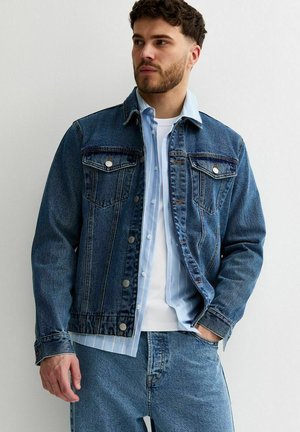 Hombre con chaqueta de mezclilla azul en capas, camisa de rayas azul claro y camiseta blanca, de pie con una mano en el bolsillo contra una pared blanca.