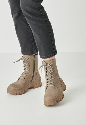 Beige vetercombatboots met zijritsen gedragen met zwarte cropped broeken op een lichte achtergrond.
