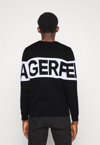 Musta neulepusero, jossa valkoinen nauha mustilla kirjaimilla, jotka muodostavat sanan "AGERFELD." Resoroidut hihansuut ja helma, klassinen pyöreä pääntie.