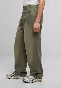 Pantalones de pierna ancha color oliva hechos de algodón. Presentan lazos para cinturón, bolsillos delanteros y un diseño clásico de cinco bolsillos, combinados con zapatillas de correr plateadas.