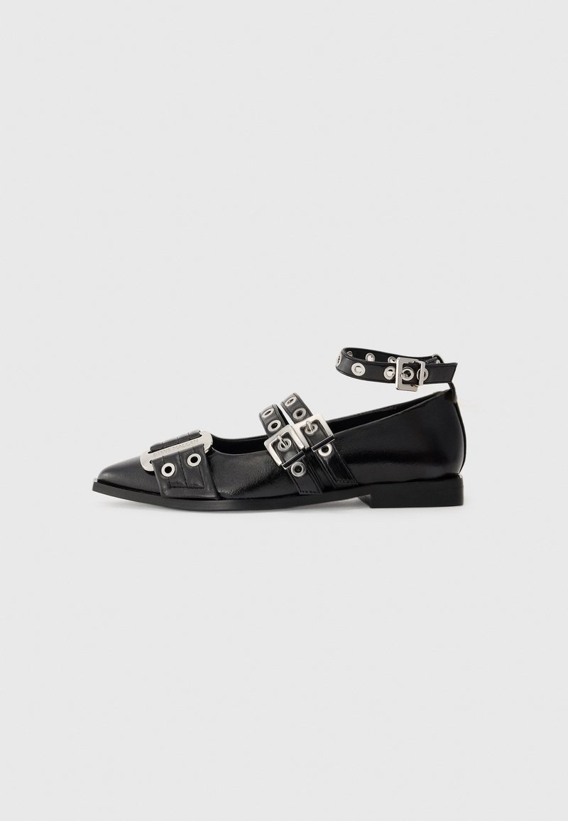 Chaussure plate noire à bout pointu avec trois brides à boucles argentées et bride de cheville avec œillets argentés sur fond clair uni.