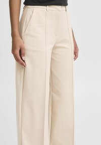 Beige weite Hosen mit Bundfalten, Gürtelschlaufen und Seitentaschen. Glatte Stofftextur. Entworfen für eine lockere Passform.