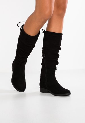 Jambes portant des bottes mi-mollet en daim noir, talons bas, fermeture éclair latérale et cordons de serrage en haut, sur fond blanc.