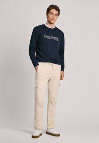 Marineblauwe sweatshirt met ronde hals en wit "Pepe Jeans" logo, gecombineerd met ivoorkleurige cargobroek met zijzakken en crèmekleurige sneakers.