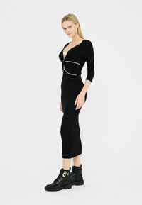 Una donna posa con sicurezza in un vestito nero aderente con scollatura profonda e dettagli di zip dorata, abbinato a stivaletti neri con lacci.