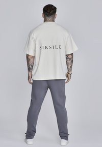 Krémové oversized tričko s čiernym logom "SIKSILK" na chrbte, v kombinácii s šedými teplákmi a bielymi teniskami. Textúrovaná bavlnená tkanina.