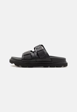 Sort åben tå slide sandal med to justerbare spænderem og tyk mønstret sål, mærket med præget UGG-logo på den øverste rem.