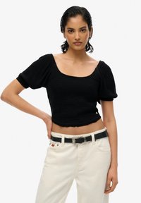 Kvinde med slicked-back hår iført en sort puff-ærmet crop top og hvide bukser med et sort bælte, stående foran en ensfarvet baggrund.