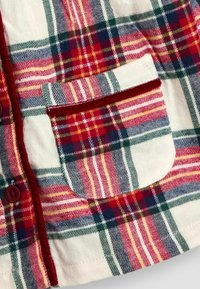 Karierte Flanellhemd in Rot, Grün und Gelb mit einer Brusttasche. Weiche Textur, Knopfverschluss und kontrastierendes rotes Bändchen entlang der Knopfleiste.