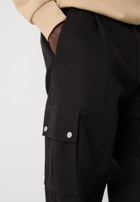 Black cargo pants with a side pocket featuring snap closures, made of textured fabric. The waistband is elastic with a fitted design.