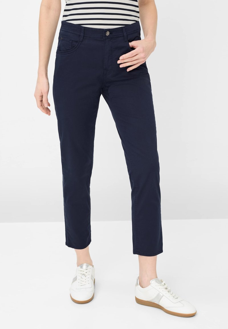 Person iført marineblå, slim-fit bukser, hvide sneakers med sølvdetaljer og en hvid og marineblå stribet skjorte, stående med den ene hånd på hoften.