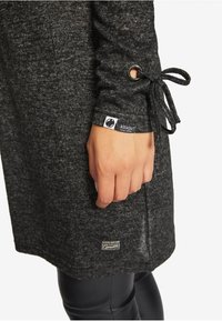 Langes, dunkelgraues Kleidungsstück mit strukturiertem Finish. Verfügt über einen gebundenen Ärmel, Ösen-Detail und ein Markenetikett nahe dem Saum.