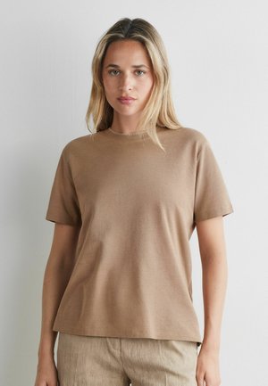 Ung kvinde med blondt hår iført en ensfarvet brun kortærmet t-shirt og beige bukser, står op ad en hvid væg.