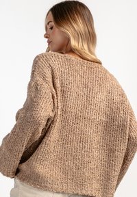 Beżowy sweter dzianinowy z teksturowanym wzorem. Posiada luźny krój, opadające ramiona i prążkowany dół. Pokazany widok z tyłu.