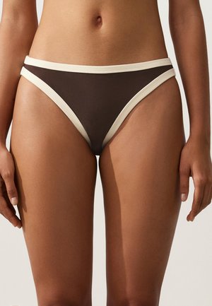 Frau trägt braune Bikinihose mit weißem Rand, steht mit entspannten Armen an den Seiten vor einem einfarbigen Hintergrund.