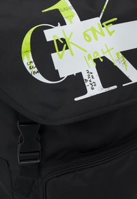 Mochila de tela negra con solapa, que presenta gráficos de logotipo en blanco y verde neón, así como múltiples detalles de texto manuscrito. Cierre de hebilla ajustable.