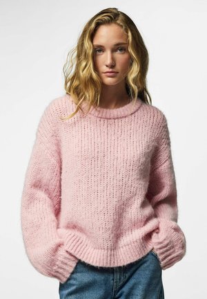Jeune femme aux cheveux blonds ondulés portant un pull en tricot rose doux et un jean bleu, mains dans les poches, expression neutre, fond blanc.