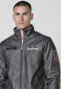 Chaqueta de motocicleta de cuero gris con cuello alto, cierre de cremallera, parches con logotipos y costuras en contraste. Cuenta con detalles bordados en las mangas.