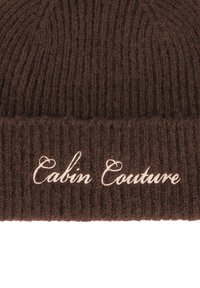 Brun ribbestrikket lue med brettet kant, med brodert tekst "Cabin Couture" i lys beige. Teksturert ullmateriale.