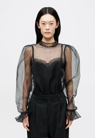 Blouse noire en voile avec des manches bouffantes, des détails en dentelle florale et des accents en cristal au niveau du col, assortie à un pantalon noir ajusté.