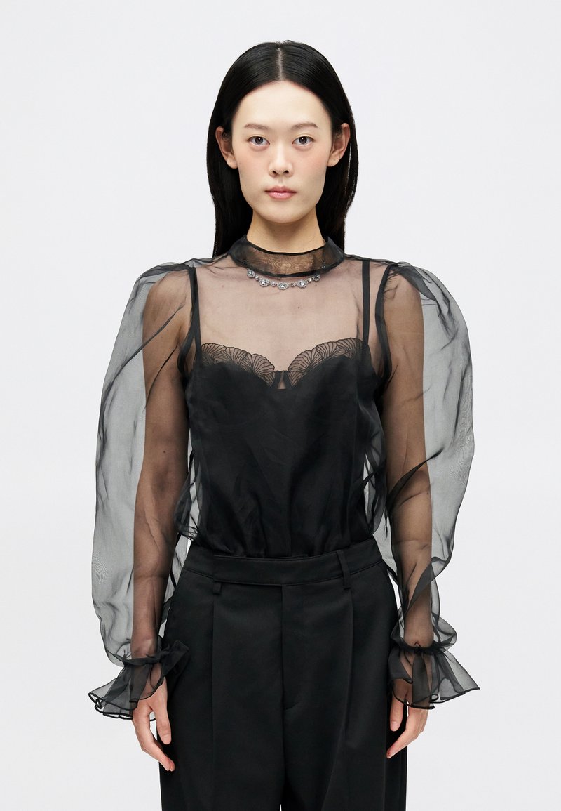 Μαύρη διάφανη μπλούζα με φουσκωτά μανίκια, λουλουδάτα δαντελένια διακοσμητικά και κρυστάλλινες λεπτομέρειες στον λαιμό, συνδυασμένη με κομμένα μαύρα παντελόνια.