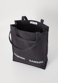 Black canvas tote bag with dual handles, featuring a simple design and a white logo text "SAM SøE" on the front. Open top with interior.