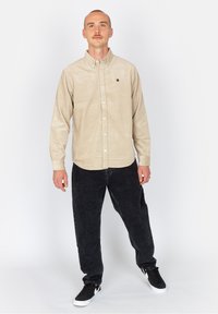 Camisa de botones de pana beige con cuello, que presenta un pequeño logo. Combinada con pantalones negros de corte holgado y zapatillas negras.