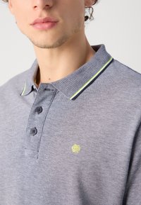 Camisa polo gris de tela texturizada, con una tapeta de dos botones, un cuello con estampado y un pequeño logotipo amarillo en el pecho.