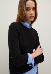Suéter negro de punto con un escote redondo sobre una camisa de rayas azul claro. El modelo tiene los brazos cruzados, mostrando los detalles de los puños.