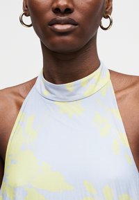 Κοντινό πλάνο προσώπου που φορά χρυσούς κρίκους στα αυτιά και ένα αμάνικο, στενό στο λαιμό τοπ με ανοιχτό μπλε και κίτρινα λουλουδένια σχέδια.