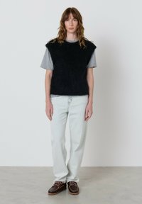 Moteris su banguotais rudais plaukais stovi vilkėdama juodą pūkuotą be rankovių megztinį virš šviesiai pilkos marškinėlio, šviesiai mėlynus džinsus ir rudus batus patalpoje.