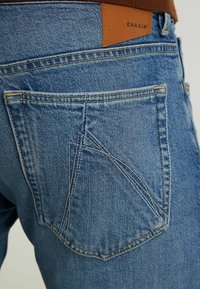 Närbild av bakfickan på blå denimjeans med en distinkt sydd design och en brun lädermärkeslapp.