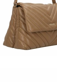 Sac à main beige matelassé avec rabat à pliage, présentant des coutures diagonales et un logo doré subtil. Matière synthétique lisse, forme structurée.